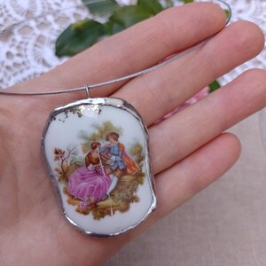 May include: A white porcelain pendant with a silver trim, featuring a painted image of a couple in a garden setting. The pendant is attached to a silver chain.