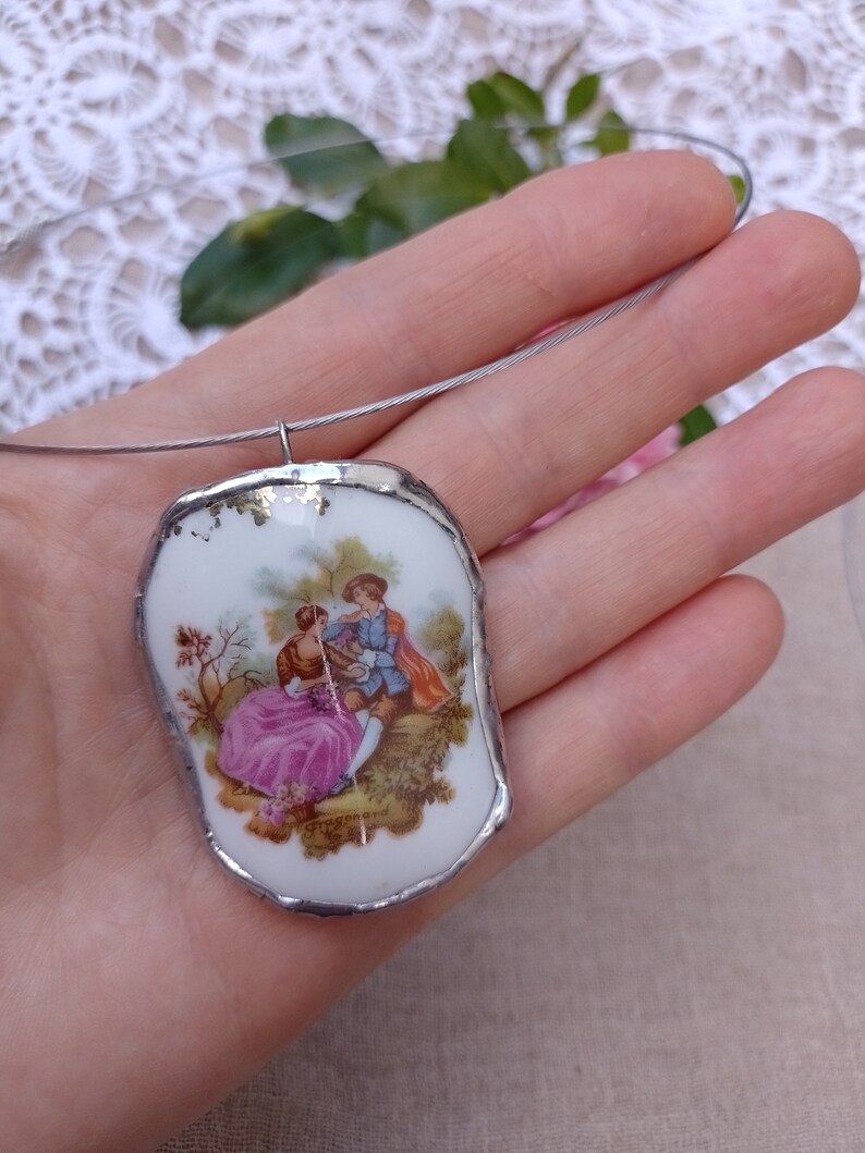 May include: A white porcelain pendant with a silver trim, featuring a painted image of a couple in a garden setting. The pendant is attached to a silver chain.