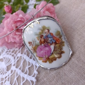 May include: A silver chain necklace with a porcelain pendant featuring a painted scene of a couple in a garden. The pendant has a silver rim and gold accents.