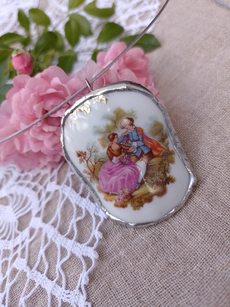 May include: A silver chain necklace with a porcelain pendant featuring a painted scene of a couple in a garden. The pendant has a silver rim and gold accents.