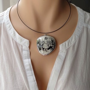 Pu&ograve; includere: Una collana con cordoncino nero con un ciondolo ovale bianco che presenta un'immagine in bianco e nero di una ragazza in un giardino.