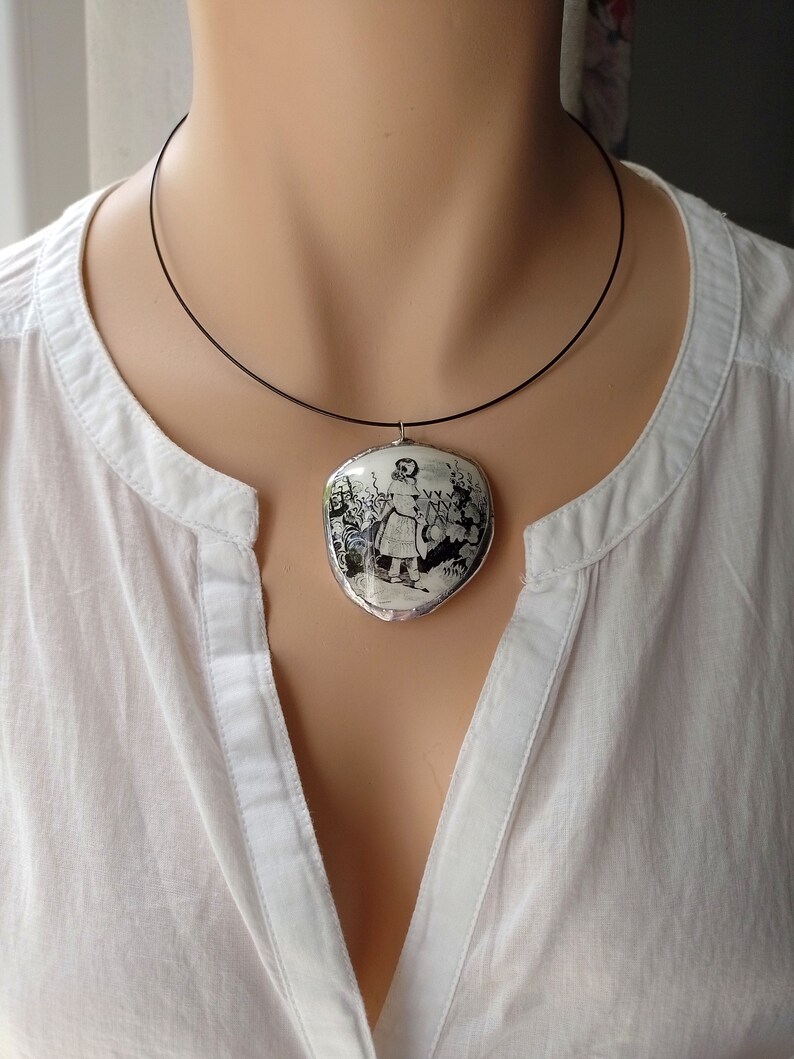 Pu&ograve; includere: Una collana con cordoncino nero con un ciondolo ovale bianco che presenta un'immagine in bianco e nero di una ragazza in un giardino.