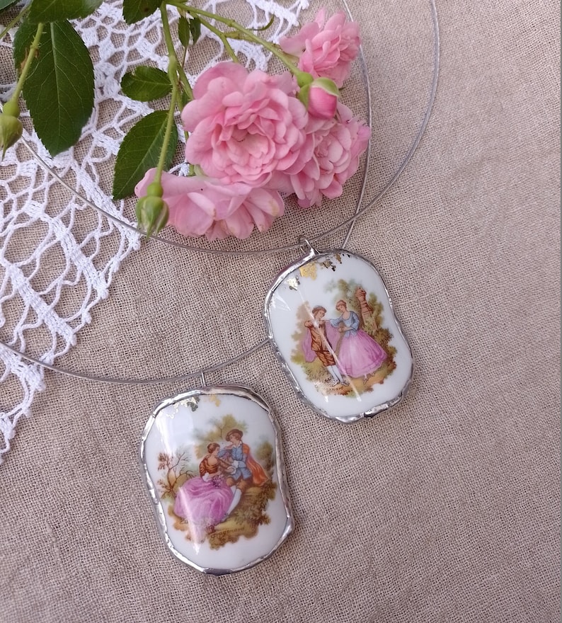 May include: A silver necklace with two porcelain pendants featuring vintage illustrations of couples in a garden setting. The pendants have gold accents and are attached to a thin silver chain.