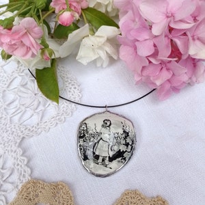 Pu&ograve; includere: Una collana con un ciondolo d'argento che raffigura un'illustrazione in bianco e nero di Alice nel Paese delle Meraviglie. Il ciondolo &egrave; appeso a un cordoncino nero ed &egrave; circondato da pizzo bianco e fiori rosa.