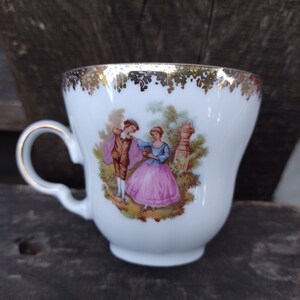 May include: A white porcelain teacup with a gold rim and a painted scene of a couple in a garden. The cup has a delicate handle.