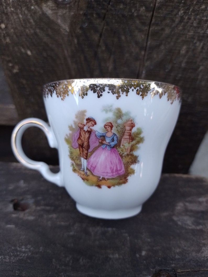 May include: A white porcelain teacup with a gold rim and a painted scene of a couple in a garden. The cup has a delicate handle.