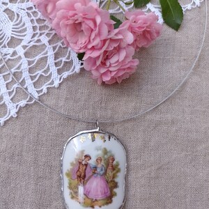 May include: A white porcelain pendant necklace with a gold trim and a vintage image of a couple in a garden setting. The necklace is on a clear cord and is surrounded by pink roses.