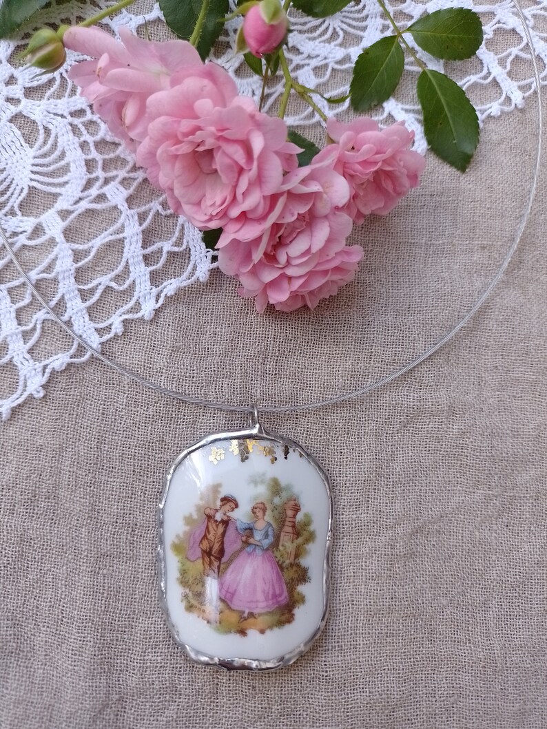 May include: A white porcelain pendant necklace with a gold trim and a vintage image of a couple in a garden setting. The necklace is on a clear cord and is surrounded by pink roses.