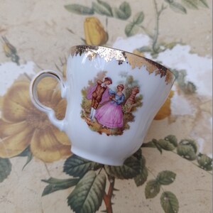 May include: A white porcelain teacup with a gold rim and a painted scene of a couple in a garden. The cup has a handle and is decorated with a gold floral pattern.