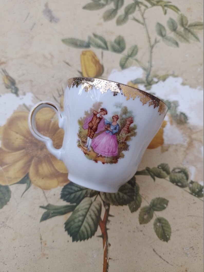 May include: A white porcelain teacup with a gold rim and a painted scene of a couple in a garden. The cup has a handle and is decorated with a gold floral pattern.