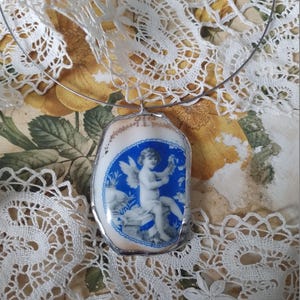 May include: A silver necklace with a blue and white porcelain pendant featuring a cherub with wings holding a flower. The pendant is shaped like a shell.