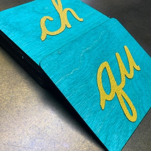 May include: A stack of teal wooden squares with rounded corners. Each square has a gold-colored cursive letter on it. The letters are "ch" and "qu". The wood grain is visible on the teal surface.