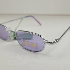 May include: Silver-framed sunglasses with rectangular, lavender-tinted lenses. The temples are silver with purple tips. The lenses have a UV 400 protection sticker and a CE mark.