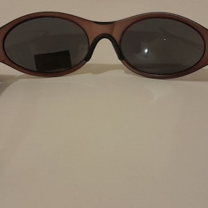 Puede incluir: Gafas de sol de pl&aacute;stico marr&oacute;n con lentes grises oscuros. Las gafas tienen un marco curvo y brazos negros.