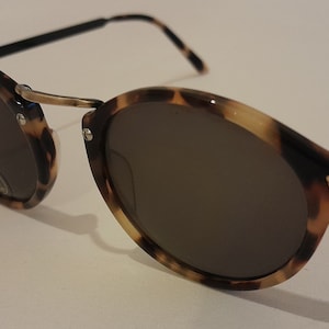 Puede incluir: Gafas de sol redondas de carey con lentes marrones. Las gafas tienen un marco de metal dorado y patillas negras.