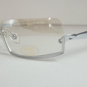 Puede incluir: Gafas sin montura con montura de metal plateado y lentes transparentes. Las gafas tienen forma rectangular y una pequeña pegatina redonda en la lente. Las patillas son de plástico transparente.