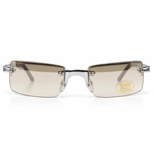 Può includere: Occhiali da sole rettangolari senza montatura con una struttura in metallo color argento lucido e lenti marroni sfumate. Un piccolo adesivo ovale sulla lente destra riporta il testo UV400 ULTRAVIOLET PROTECTION in lettere dorate su sfondo giallo.