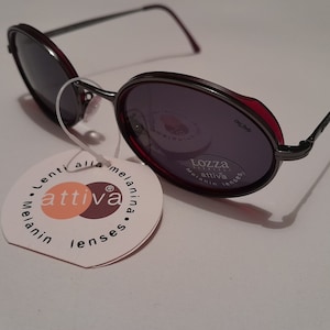 Op de afbeelding: Ronde, donkergrijze zonnebril met rode accenten op het montuur. De glazen zijn donkergrijs en hebben de tekst "Lozza" en "attiva" erop gedrukt. De zonnebril wordt geleverd met een wit label met de tekst "Lenti alla melanina. attiva. melanin lenses."