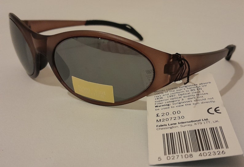 Puede incluir: Gafas de sol ovaladas de color marr&oacute;n con lentes tintadas oscuras. Las gafas de sol tienen una etiqueta negra adjunta con el texto "Kondo UV400 sunglasses absorb 100% of the sun's harmful UV rays and conform to BS EN 1836 1997. Optical class 1 superior optical quality lenses. Filter category 3 dark tint. Warning: sunglasses should not be used to view the sun directly. &pound;20.00 M207230 CE Fabris Lane International Ltd. Chessington, Surrey, KT9 1TT, UK" y un c&oacute;digo de barras.