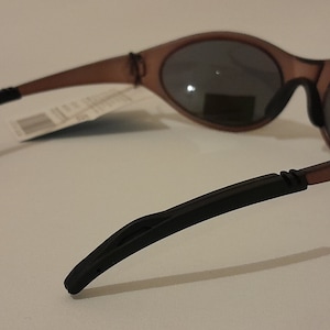 Puede incluir: Gafas de sol de pl&aacute;stico marr&oacute;n con patillas de goma negra. Las gafas tienen una forma ovalada redondeada y lentes oscuras.