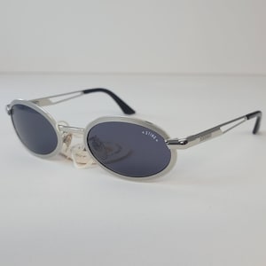 Peut inclure: Lunettes de soleil à monture argentée avec verres ovales et teintés foncés. Les lunettes ont une fine monture argentée et des branches noires. La marque "STING" est imprimée sur la lentille gauche. Lunettes neuves avec étiquette.