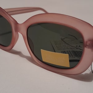 May include: Pink oval-shaped sunglasses with dark lenses. The sunglasses have a matte finish and a small tag attached to the right arm.