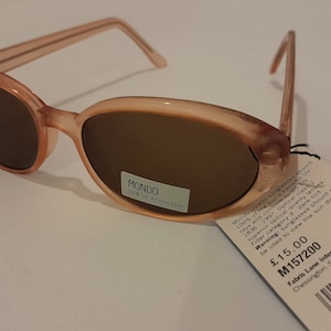 May include: Pink oval-shaped sunglasses with brown lenses. The sunglasses have a Mondo label that says "100% UV Protection". The tag attached to the sunglasses says "Mondo Sunglasses", "100% of the sun's harmful rays and 100% UV protection", "Optical Class 3", "Superior optical quality lenses", "Filter category 3 dark tint", "Warning: Sunglasses should not be used to view the sun directly", "£15.00", "M157200", "Fabris Lane International Ltd.", "Chessington, Surrey, KT9 1TT, UK.", and has a CE mark.
