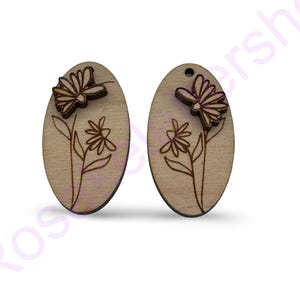 May include: A pair of wooden earrings with a floral design. The earrings are oval shaped and have a brown wood finish. The floral design is laser cut and features a butterfly and flowers.