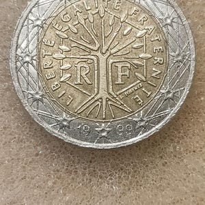 Op de afbeelding: Een munt van twee euro met een zilveren buitenring en een goudkleurig midden. De munt toont de woorden "LIBERTE EGALITE FRATERNITE" en het jaar 1999. Het midden toont een gestileerde boom.