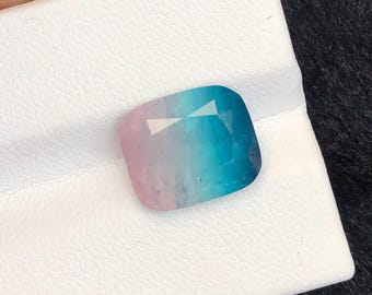 Può includere: Una gemma taglio cuscino con un gradiente di tonalità rosa, blu e turchese. La gemma è posta su una superficie bianca, con uno sfondo scuro. Le sfaccettature della gemma riflettono la luce, creando un effetto scintillante. Questo articolo è adatto per la creazione di gioielli.