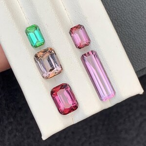 Pu&ograve; includere: Quattro pietre preziose di diversi colori, tra cui verde, rosa e viola, sono esposte su una superficie bianca. Le pietre preziose sono tutte tagliate a forma rettangolare.