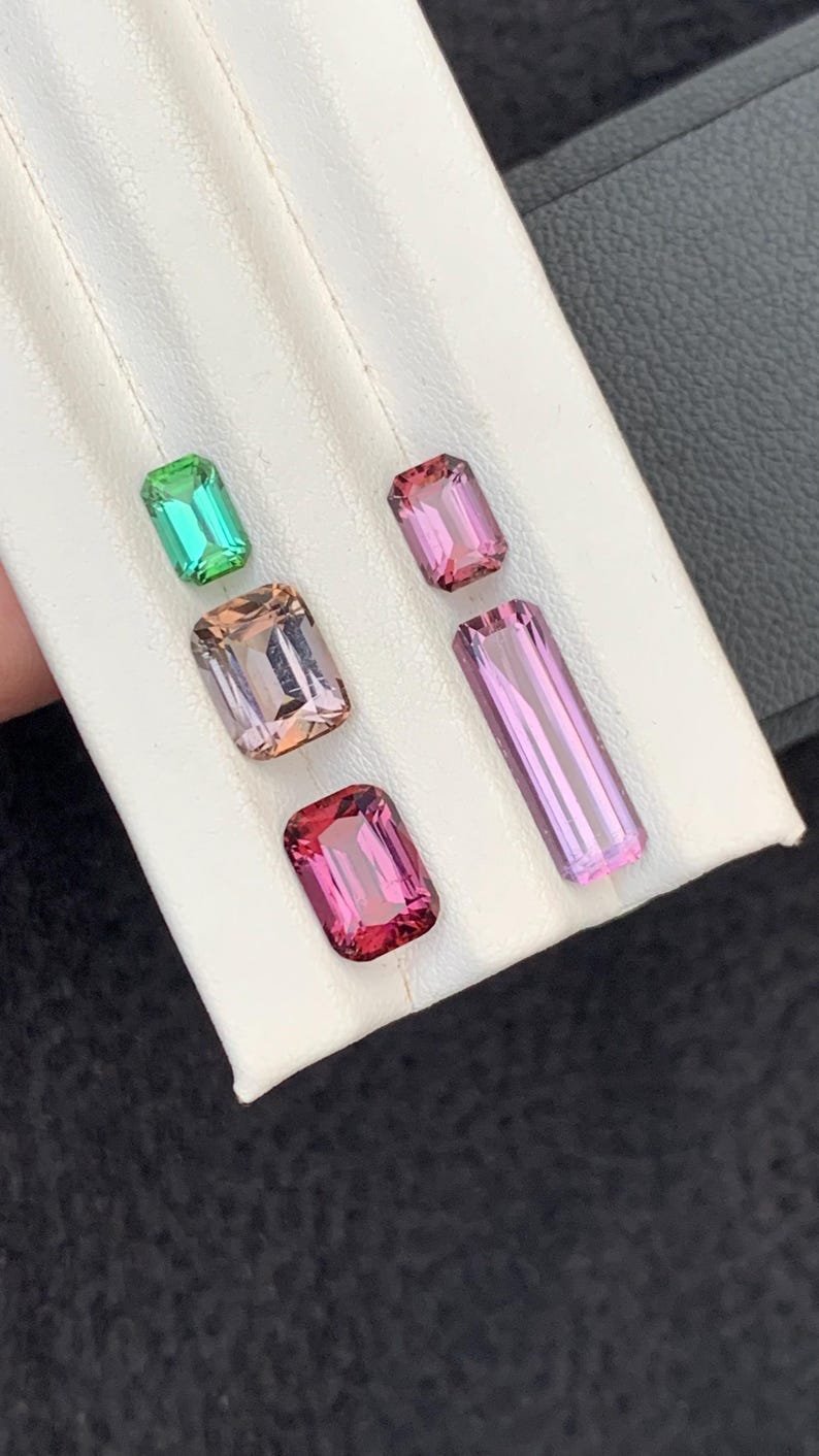 Pu&ograve; includere: Quattro pietre preziose di diversi colori, tra cui verde, rosa e viola, sono esposte su una superficie bianca. Le pietre preziose sono tutte tagliate a forma rettangolare.