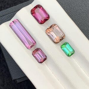 Pu&ograve; includere: Cinque pietre preziose esposte su un vassoio bianco. Le pietre preziose sono rosa, rosa chiaro, pesca e verde. Tutte le pietre preziose sono tagliate a forma rettangolare.