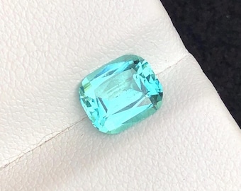 Può includere: Una pietra preziosa taglio cuscino con un vibrante colore blu ciano traslucido. La pietra presenta una superficie sfaccettata che riflette la luce, creando un aspetto brillante. Ha una forma rettangolare con angoli arrotondati e finitura lucida.