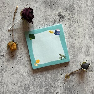Pode incluir: Um bloco de notas quadrado azul claro com ilustrações de brócolos, uma fatia de limão, manteiga e uma caixa de leite. Flores secas no fundo. A nota tem um espaço em branco para escrever.