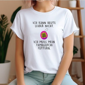 May include: White t-shirt with black text that reads "Ich kann heute leider nicht. Ich muss mein Tamagotchi füttern."  A pink and green Tamagotchi is illustrated above the text.