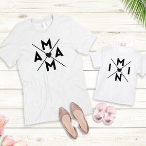 May include: Two white t-shirts with black text and a heart design. The larger shirt says "MAM" and the smaller shirt says "MINI".