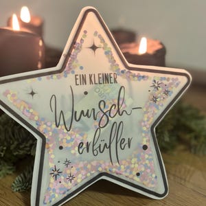 Peut inclure: Décoration en forme d'étoile avec un contour noir, remplie de confettis colorés et le texte allemand "Ein kleiner Wunscherfüller". La décoration est sur une surface en bois, avec des bougies allumées en arrière-plan.