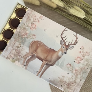 May include: A Christmas advent calendar with a deer illustration. The calendar has 24 numbered doors, each with a small chocolate behind it. The deer is standing in a snowy forest setting with pink roses.