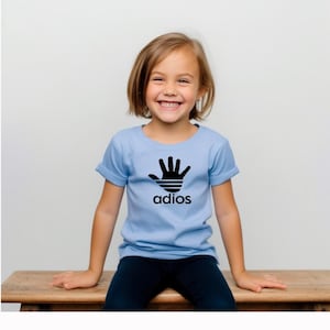 Könnte beinhalten: Hellblaues T-Shirt mit schwarzer Handgrafik und dem Wort "adios" in einer stilisierten Schriftart. Das T-Shirt wird von einer jungen Person getragen. Das T-Shirt hat kurze Ärmel und einen Rundhalsausschnitt.