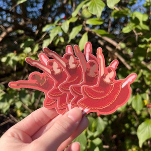 May include: Five red and gold ballerina stickers with a white outline. The ballerinas are in a variety of poses, with their arms and legs outstretched.