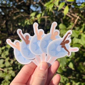 May include: Five blue and white ballerina stickers with different poses. The stickers are held in a hand against a green leafy background.