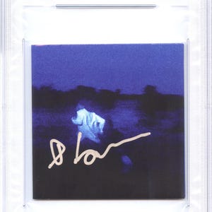 May include: A CD cover featuring a dark blue landscape with a blurred figure and a signature in white. The top of the case has a label that reads "CD COVER DANIEL CAESAR PSA/DNA CERTIFIED AUTHENTIC AUTO".