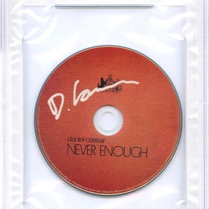 May include: A CD by Daniel Caesar, titled "Never Enough," is encased in a clear plastic holder. The orange disc features the artist's signature and the album title. A PSA/DNA Certified label is visible at the top.