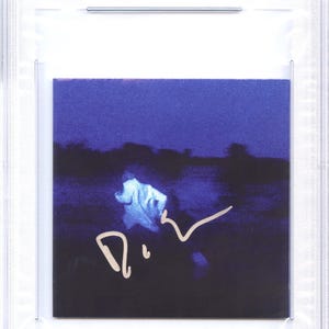May include: A CD cover featuring a dark blue image with a blurred figure and a signature in gold ink. The cover is encased in a clear plastic holder with a label that reads "CD COVER DANIEL CAESAR PSA/DNA CERTIFIED AUTHENTIC AUTO 85670731".