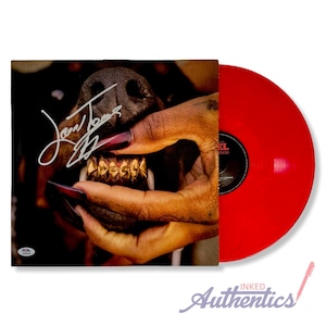 May include: A red vinyl record next to a record sleeve featuring a close-up of a dog's face with gold teeth. A hand with long, red nails is holding the dog's mouth open. The sleeve is signed.