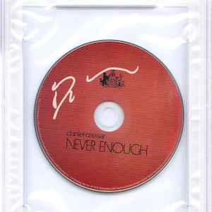 May include: A Daniel Caesar "Never Enough" CD in a protective case. The CD is red with white text and a logo. The case has a PSA/DNA certification label.