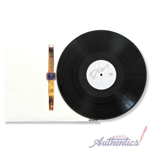 May include: A gold-toned wristwatch with a square blue face and a black vinyl record. The record has a white label with the text "Never Enough" and a signature. The items are on a white background.