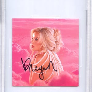 May include: A CD insert featuring Megan Moroney, PSA/DNA certified. The insert shows a woman with blonde hair and a pearl necklace against a pink cloudy background. The autograph "Megan" is visible.