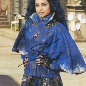 May include: A woman wearing a blue jacket with gold studs and a matching skirt. The jacket has a high collar and a zipper. She is wearing black fingerless gloves and a necklace with a red pendant. Her hair is dark blue and styled in waves.